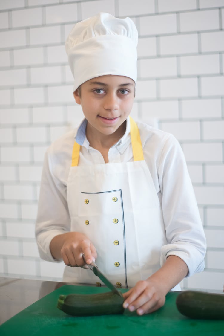 A Boy Wearing Chef's Uniform Slicing Cucumber While Looking At The Camera