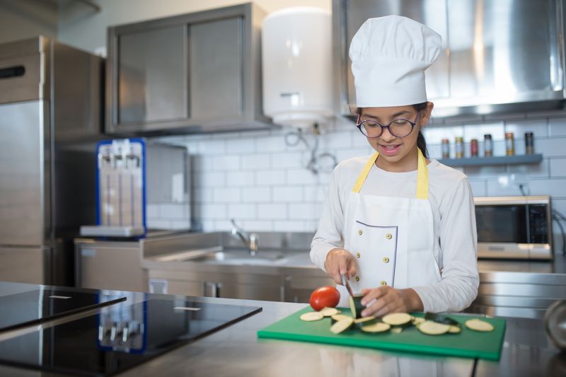 Girl in chef's hat slices vegetables on a chopping board in a modern kitchen.