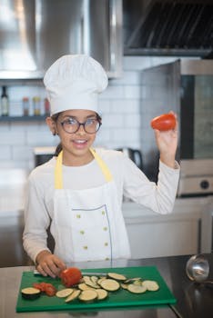 Young girl in a kitchen holding tomato, preparing zucchini slices on a chopping board.