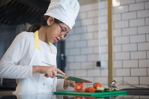 A young girl wearing a chef hat and glasses slices vegetables in a modern kitchen.