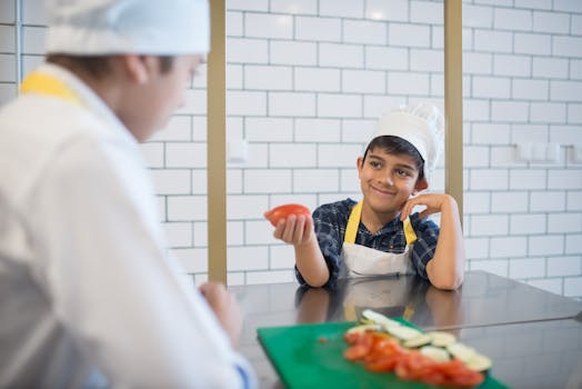 Smiling young boy wearing a chef's hat in a kitchen, proudly holding a tomato.