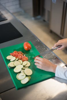 Hands slicing tomatoes and zucchini on a green cutting board in a kitchen.