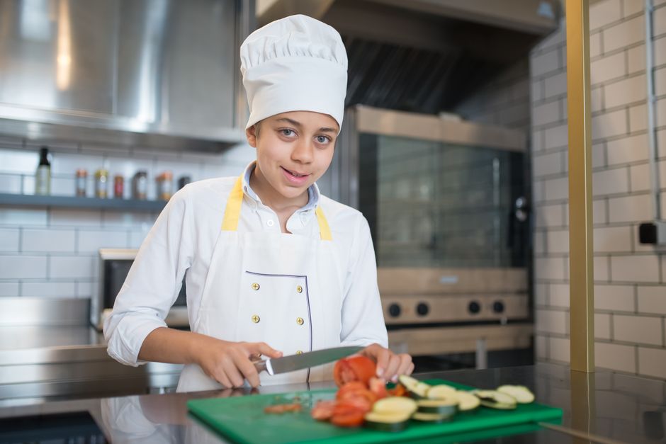 A young chef in a professional kitchen slicing vegetables on a cutting board.
