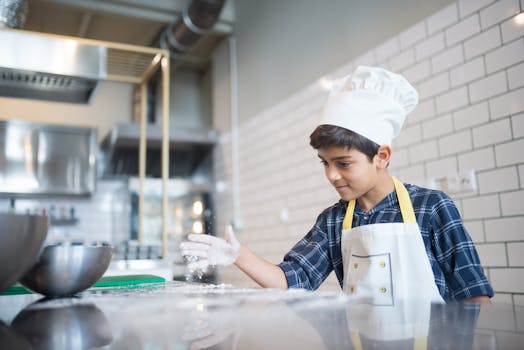 A young boy in a chef's hat and apron sprinkles flour on a kitchen countertop, showcasing his cooking skills.