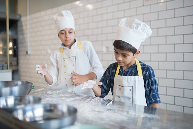 Young Kids Wearing Toque And Aprons Inside The Kitchen While Playing Flour