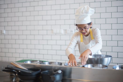 A young girl wearing a chef's hat and apron cooks enthusiastically in a modern kitchen setting.