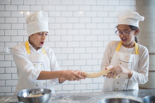 Two kids in chef hats having fun while stretching dough in a kitchen. Perfect culinary learning moment.