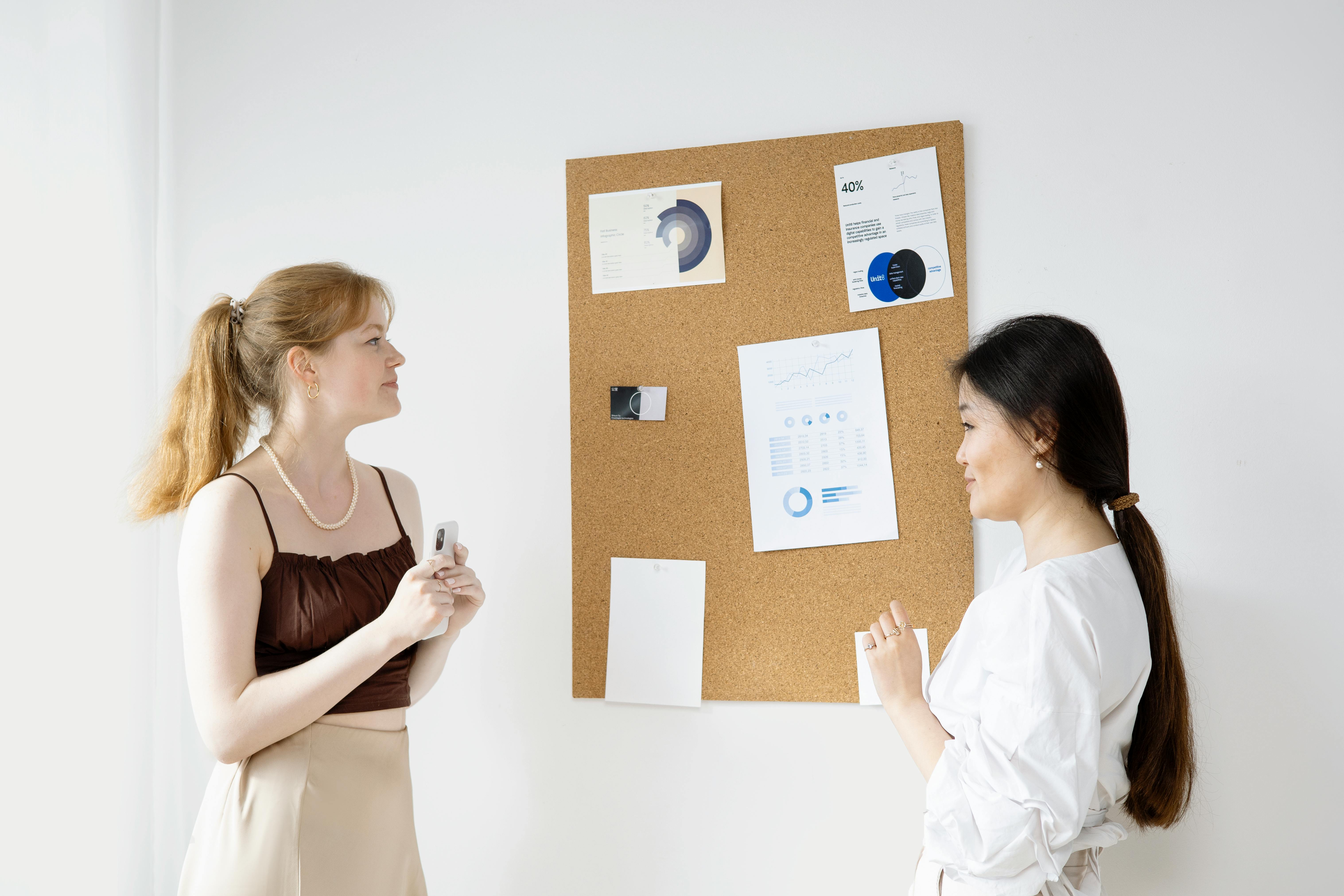Free Two women discussing charts on a bulletin board in an office setting. Stock Photo