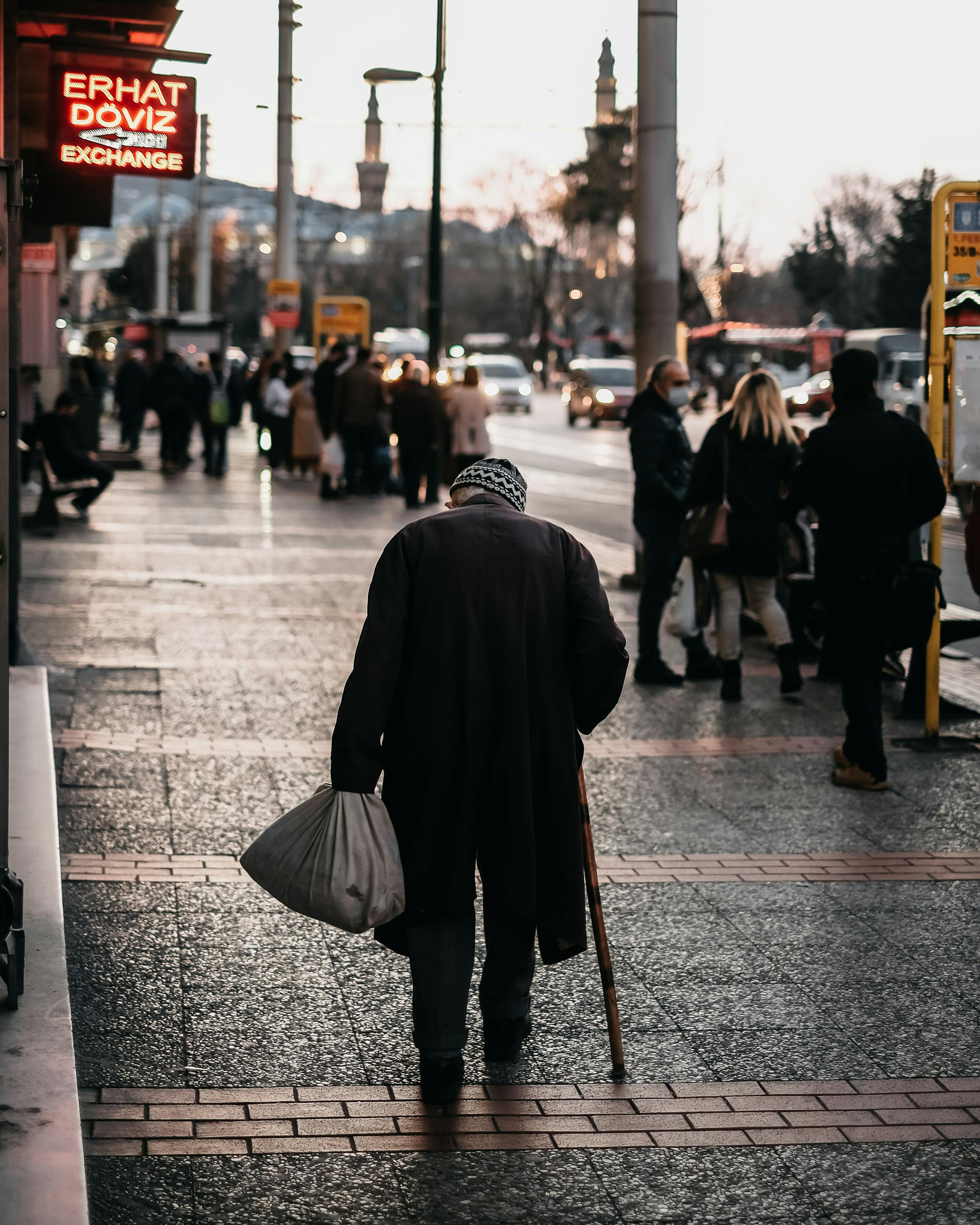 An elderly man walks on a city sidewalk at dusk, surrounded by urban life.
