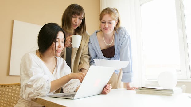 Three women in a modern office setting collaborating around a laptop and discussing documents.