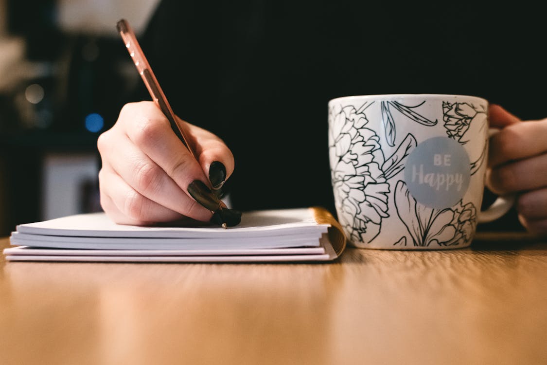 Person Writing on a Notebook near a Be Happy Painted Mug