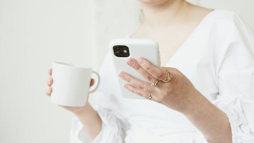 Free A Person in White Long Sleeves Holding a Cup of Coffee while Typing on Her Cellphone Stock Photo
