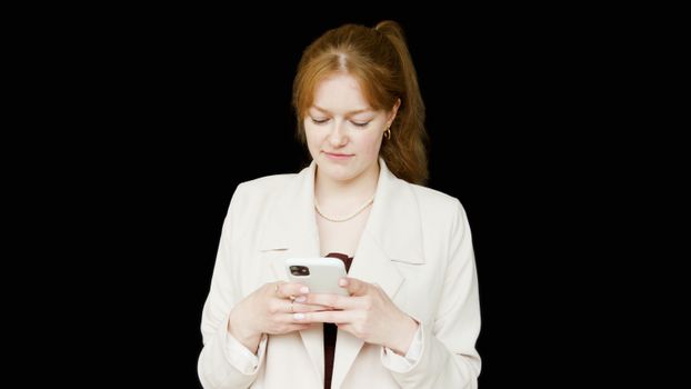 Woman in white blazer looking at smartphone against a black background, studio shoot.
