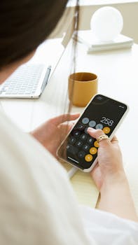 A woman calculating on a smartphone at a workspace with a laptop and mug.