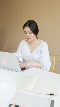 A young woman in a white blouse is focused on typing on her laptop at a desk indoors.