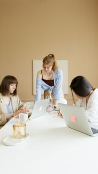 Three women collaborating in a modern office, focused on laptops and tasks at a white desk.