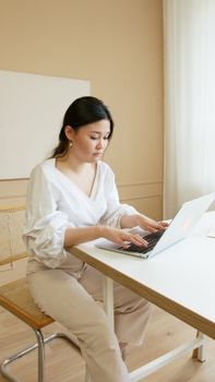 Asian woman working on a laptop in a minimalist beige office. Professional and focused workspace setting.