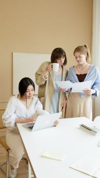 Three women collaborating in an office, using laptop and documents, drinking coffee.