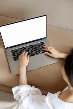 Woman sits on a couch typing on a laptop with a blank screen.
