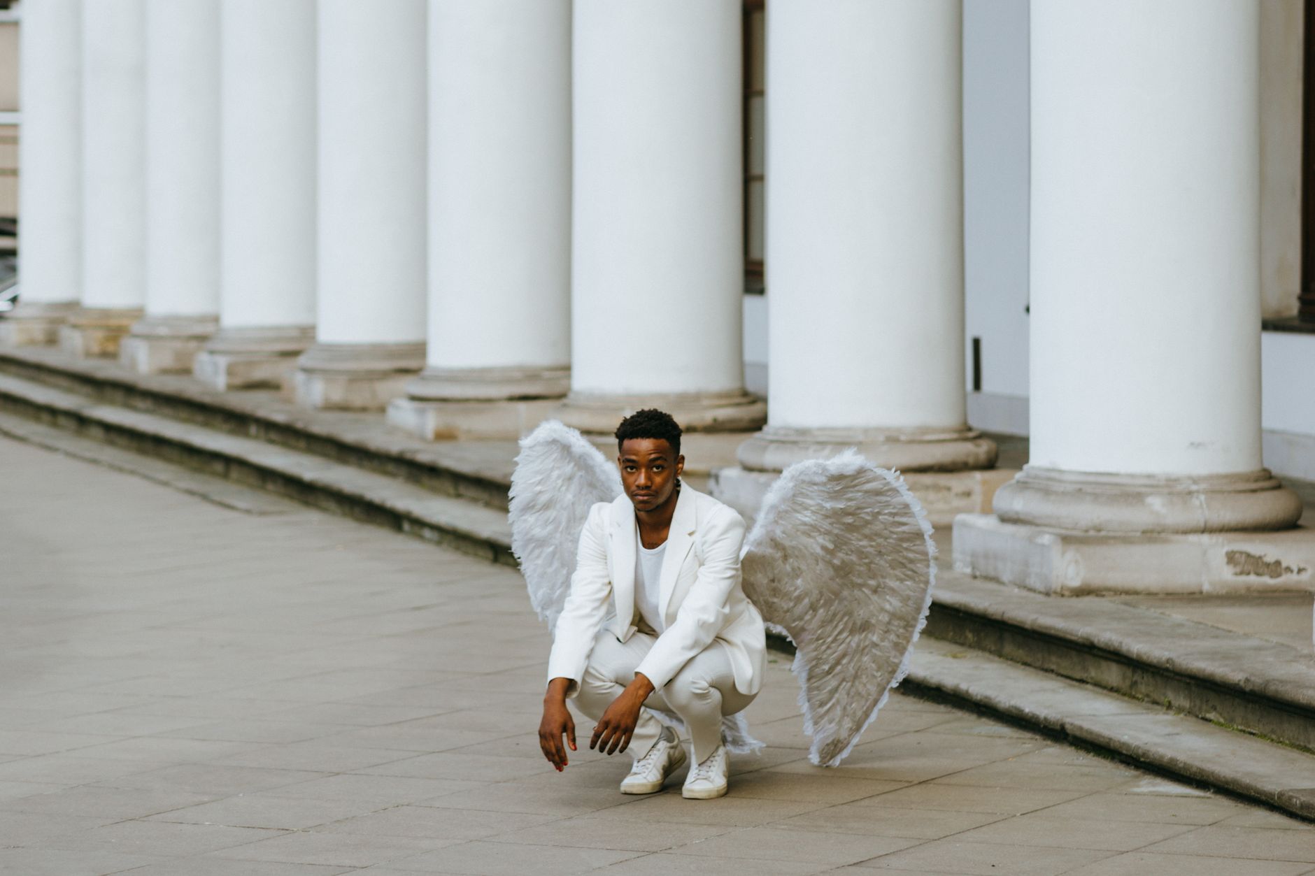 A man in a white suit with angel wings kneels elegantly outdoors against a column backdrop.