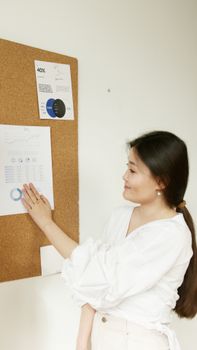 An Asian woman analyzes data charts pinned on a cork board in an indoor setting.