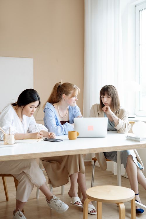 Free Three women collaborating at a desk in a bright office setting, showcasing teamwork and strategy planning. Stock Photo