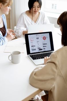 Three women discussing business strategies at a desk while showing data on a laptop in a bright office.