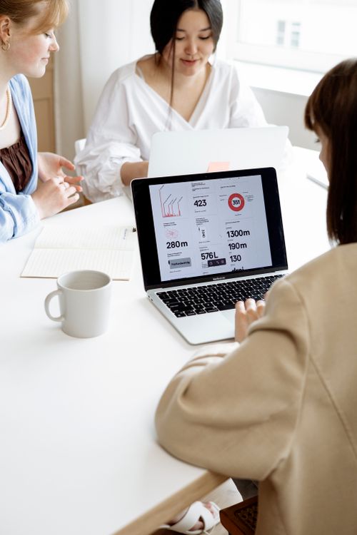 Free Three women discussing business strategies at a desk while showing data on a laptop in a bright office. Stock Photo