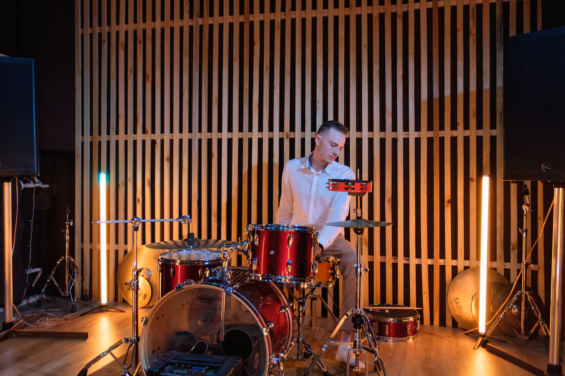 A male drummer plays a red drum set in a cozy studio with warm lighting.
