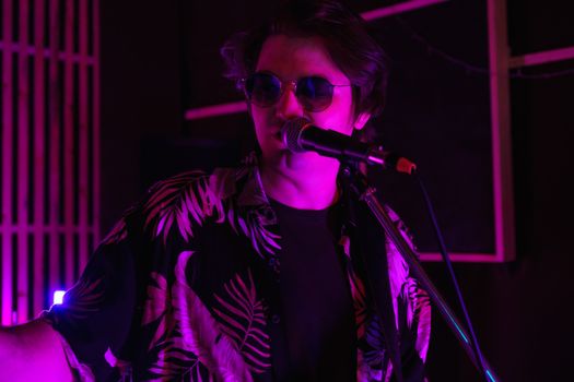 A stylish man wearing sunglasses sings into a microphone under moody purple lighting.