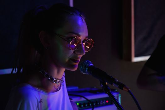 A woman vocalist wearing sunglasses and chain necklace sings into a microphone, captured in low light.