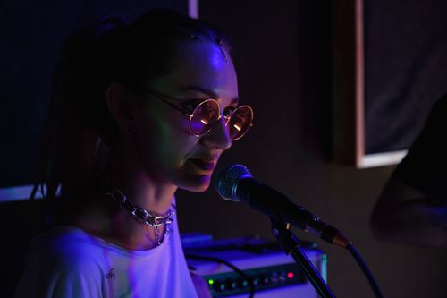 Free A woman vocalist wearing sunglasses and chain necklace sings into a microphone, captured in low light. Stock Photo