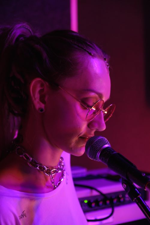 Free A woman wearing sunglasses sings into a microphone under soft purple lighting, creating a moody atmosphere. Stock Photo