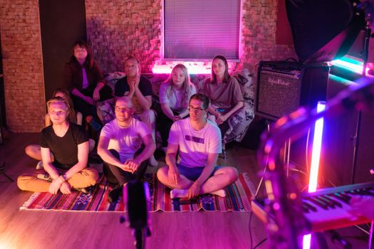 A group of people sitting indoors enjoying a home concert with neon lighting and musical instruments.