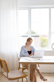 Adult woman working at a laptop in a bright, modern office setting with large windows.