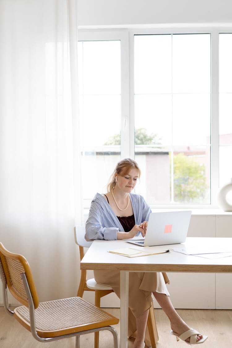 A Woman In Blue Long Sleeve Shirt Sitting On A Wooden Chair While Looking At Her Laptop