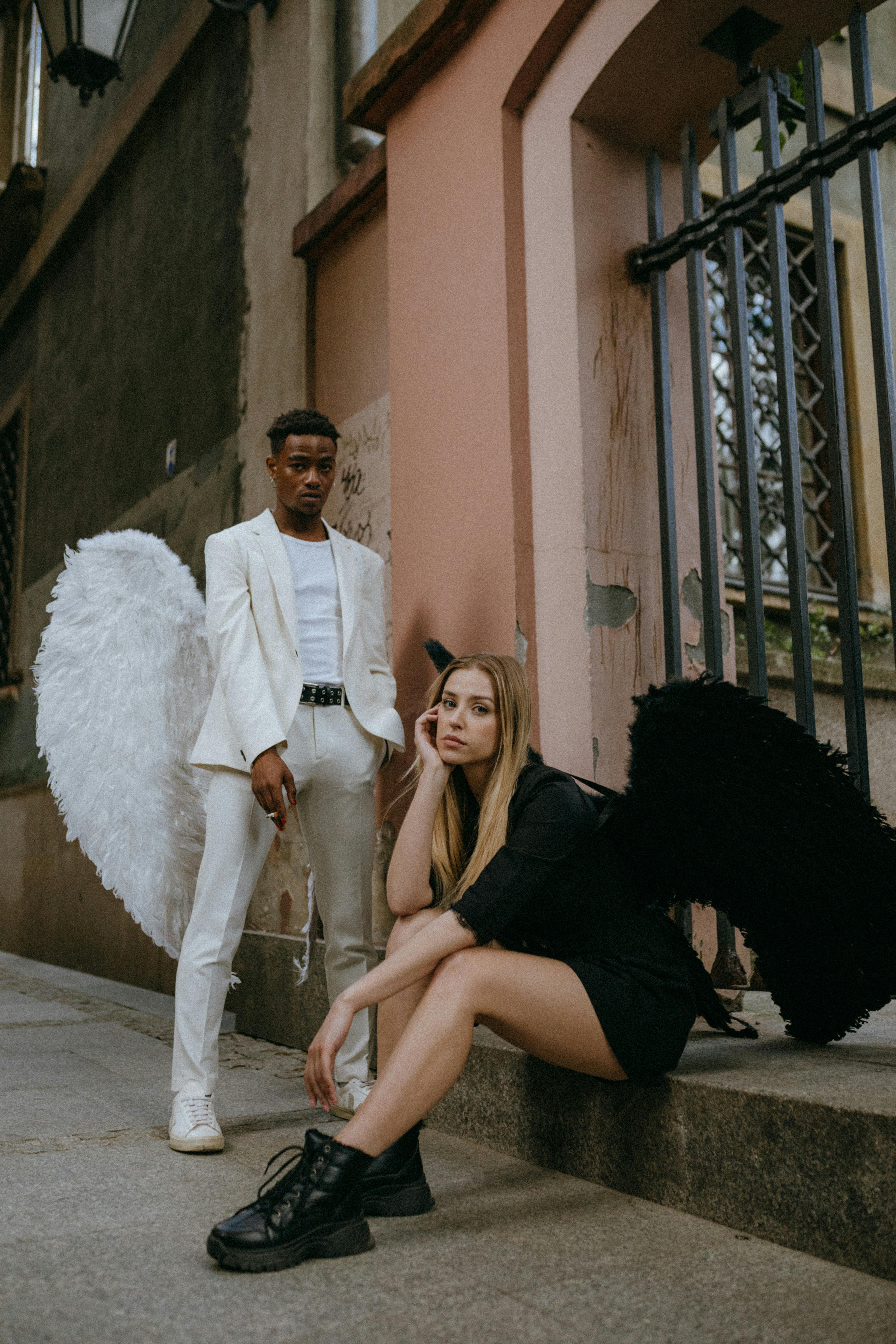 Stylish duo posed as angel and demon in an urban setting. Contrasting outfits, white and black wings.