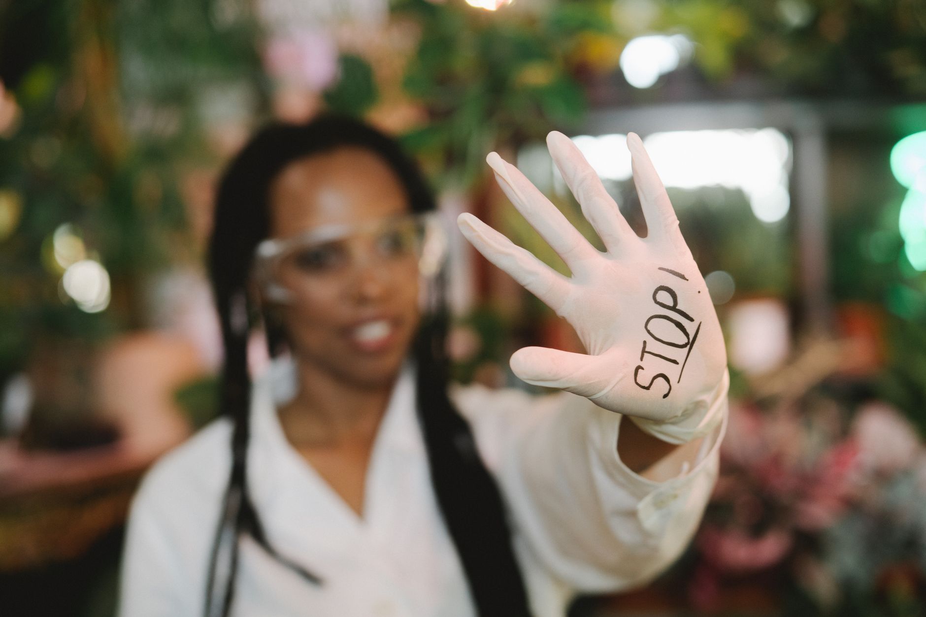 Woman holding up a hand with the word 'STOP' on a latex glove in a garden backdrop.