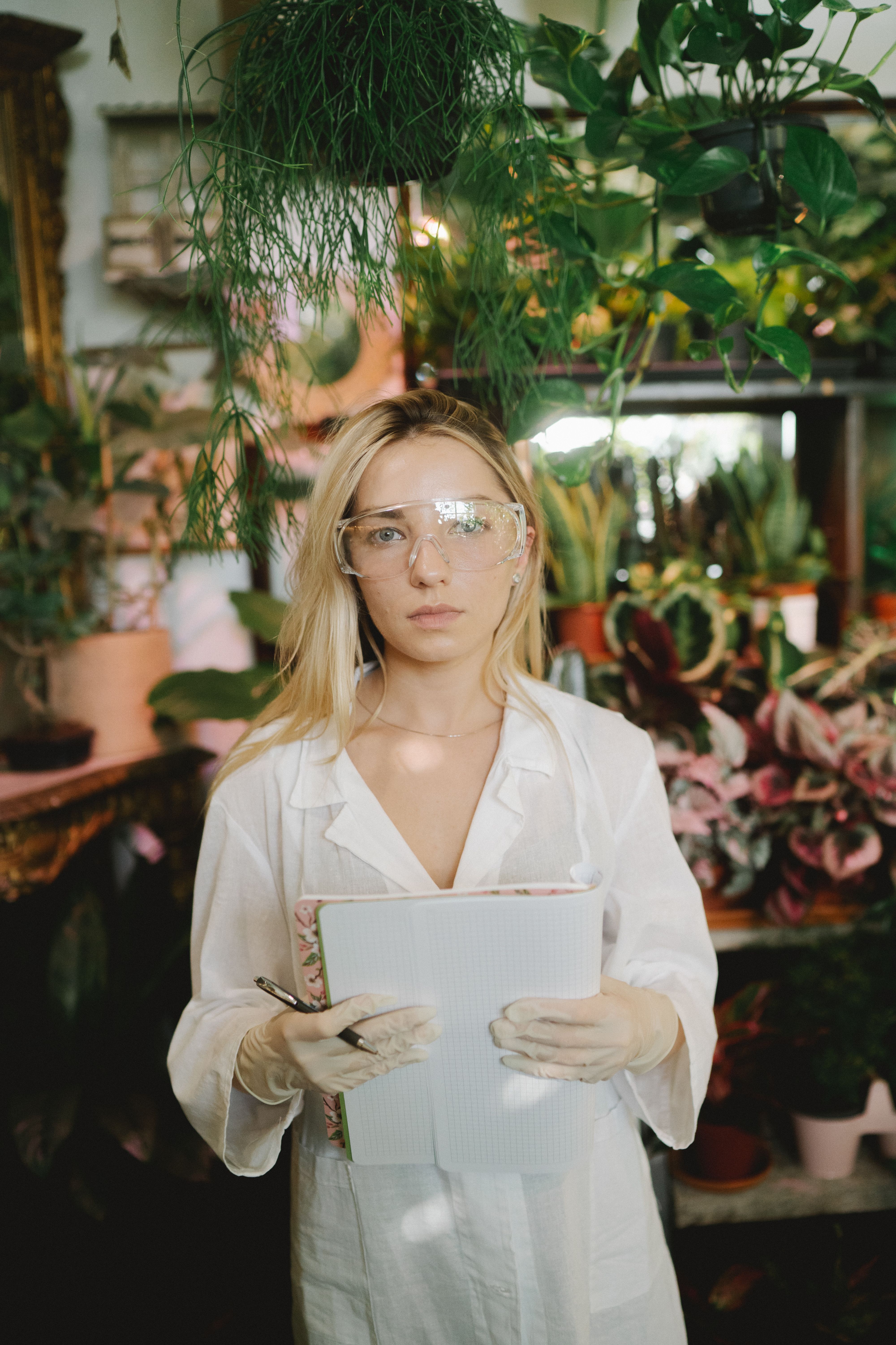 Scientist with notebook amidst plants in a greenhouse setting, wearing gloves and goggles.