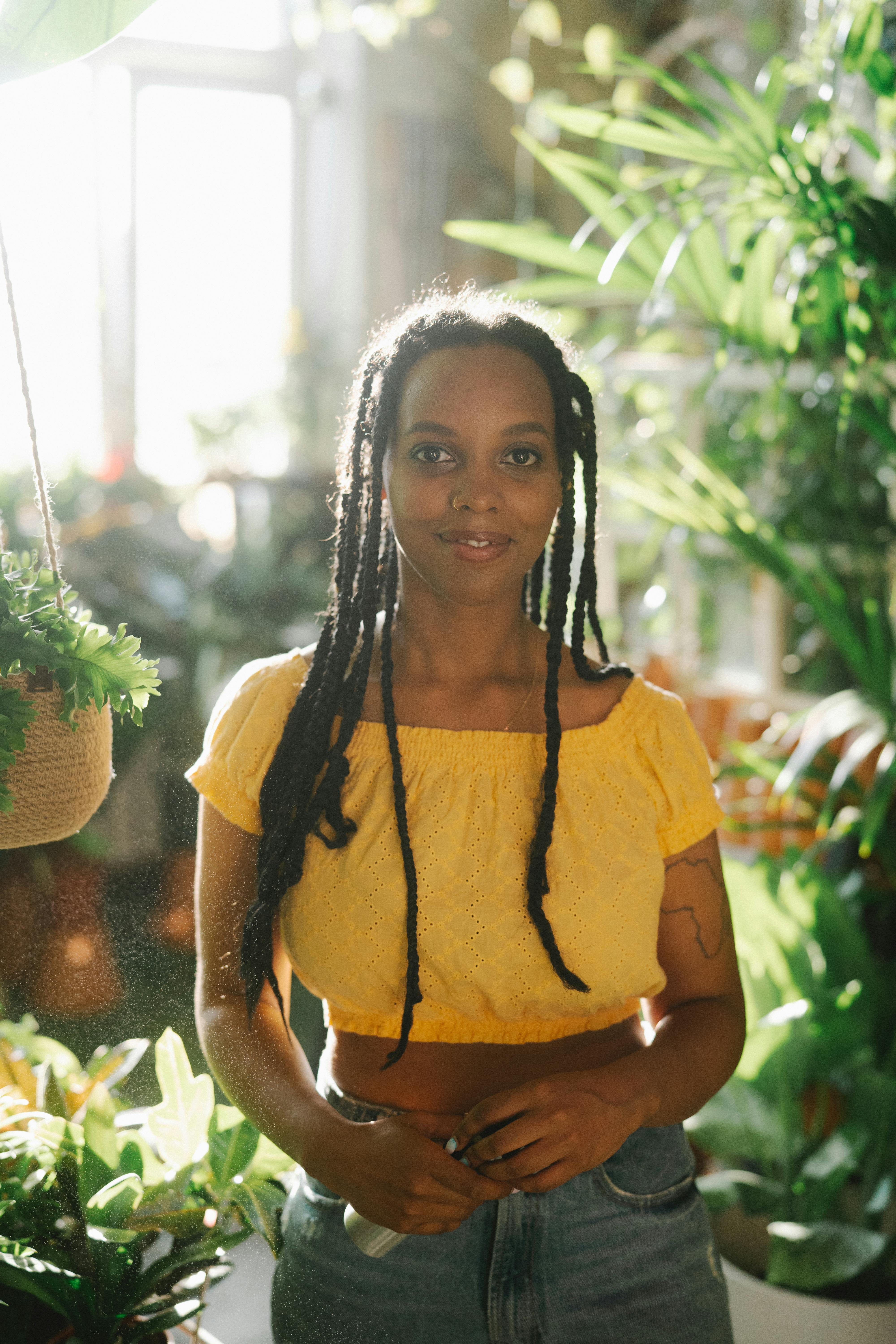 A smiling woman in a yellow top surrounded by lush indoor plants in natural daylight.