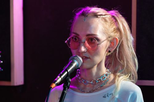 Free Portrait of a young woman singing with a microphone indoors, wearing pink sunglasses. Stock Photo
