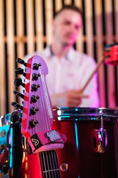 Musician blurred behind drums, guitar in focus, vibrant stage lighting.