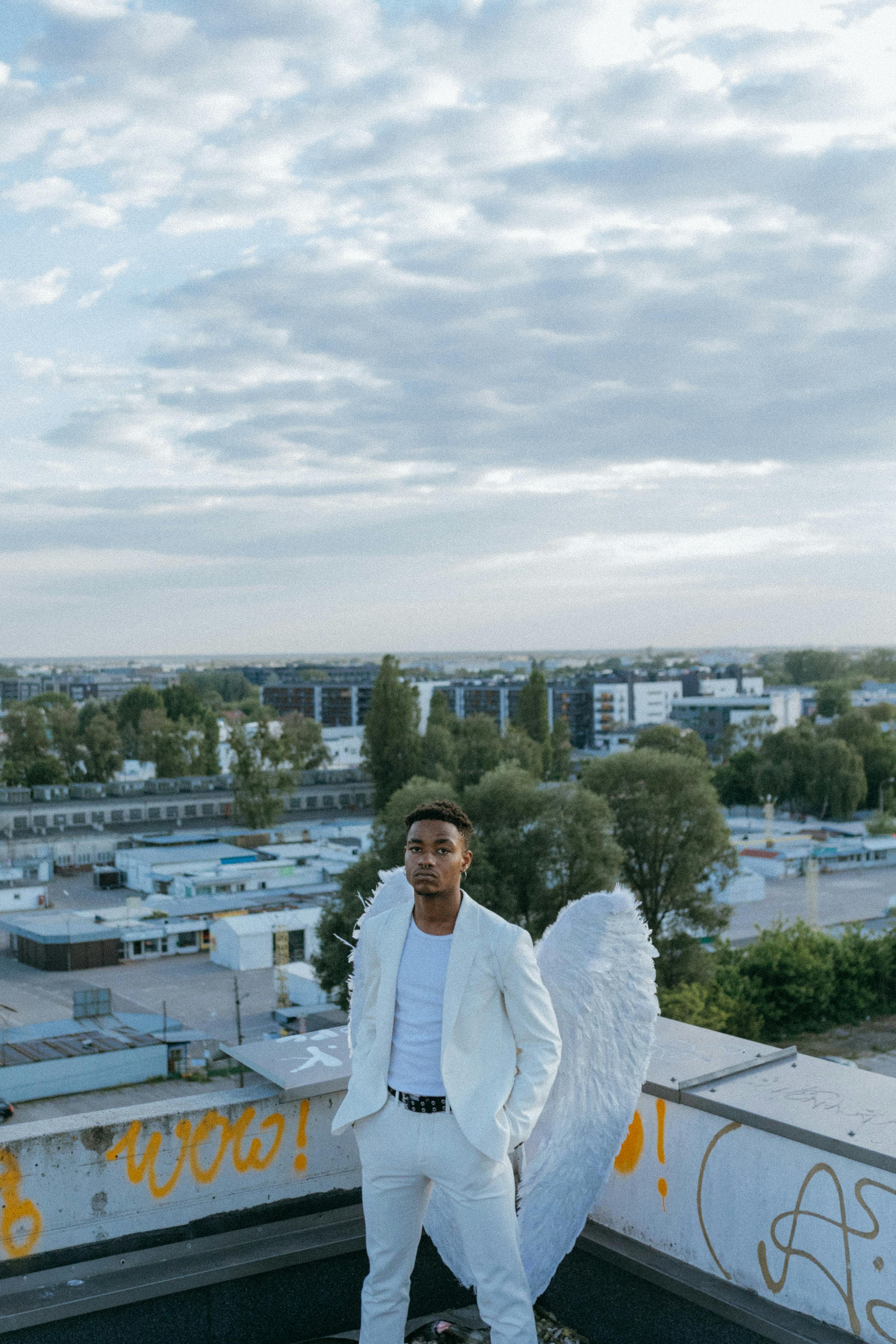 A man in a white suit with angel wings standing on an urban rooftop under a cloudy sky.