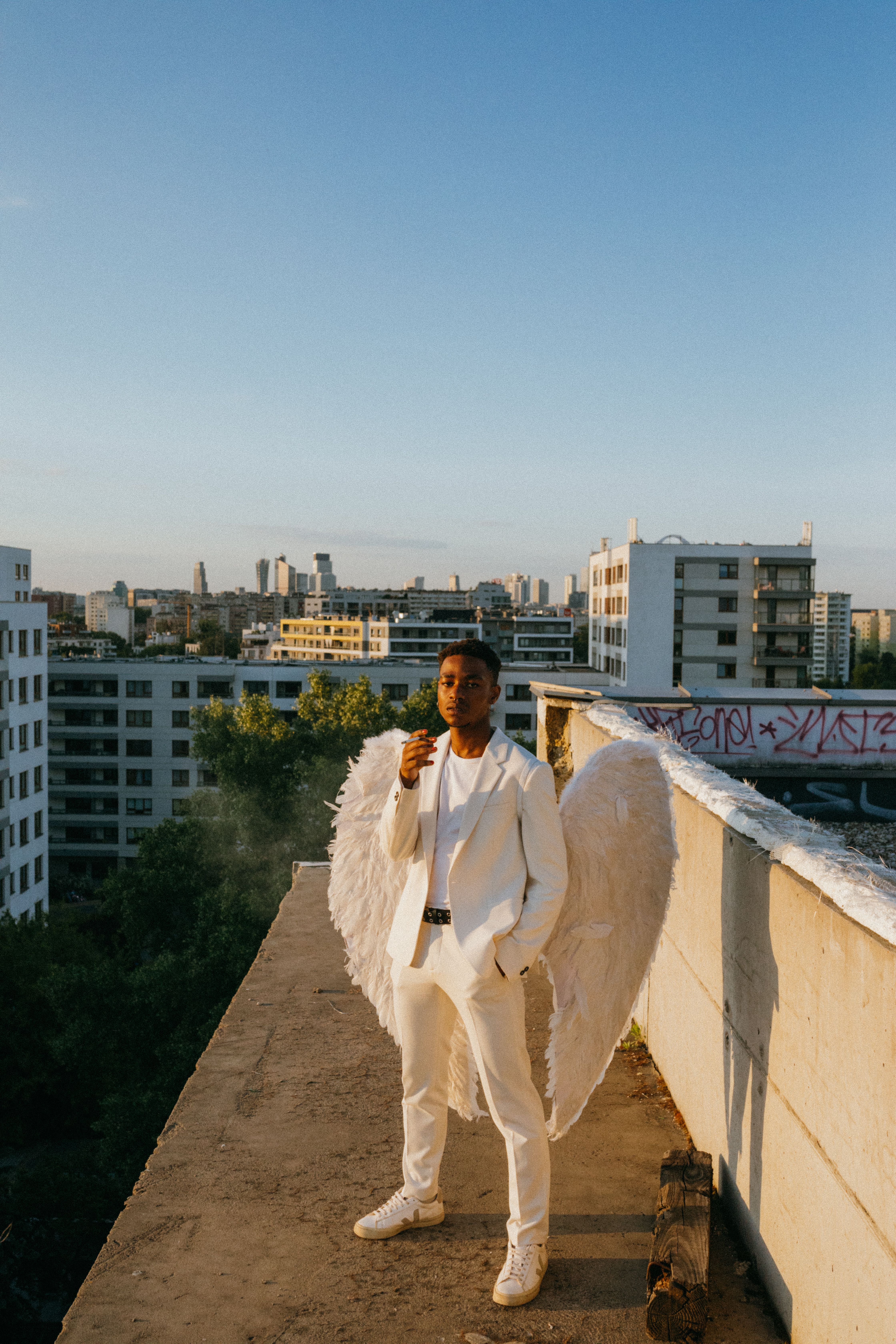 A man dressed as an angel with wings stands on a city rooftop at sunset, creating a celestial appearance.