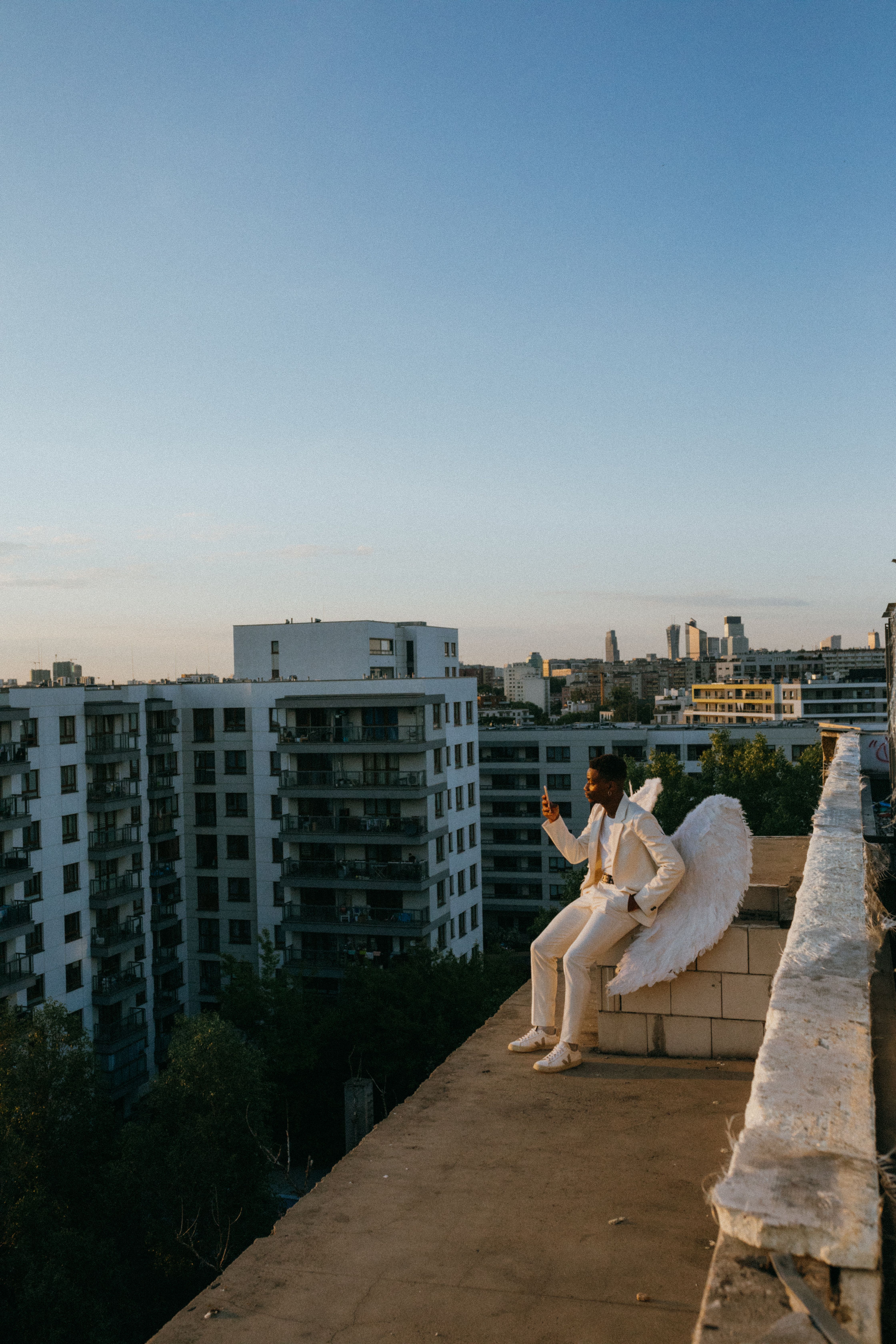A man dressed as an angel in a white suit sits on a rooftop with wings, overlooking city buildings at sunset.