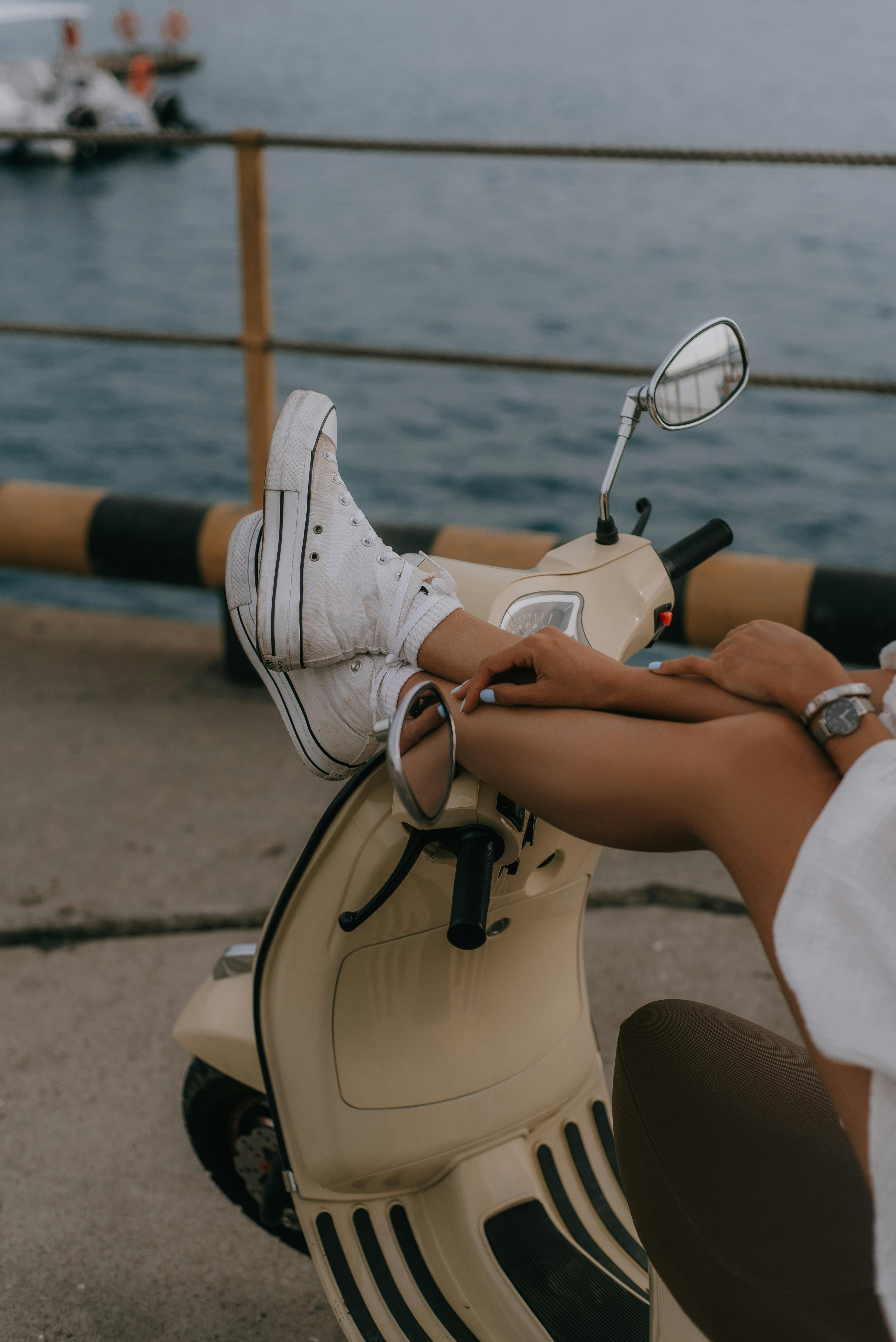Person lounging on a scooter near the sea, showcasing casual style with white sneakers.