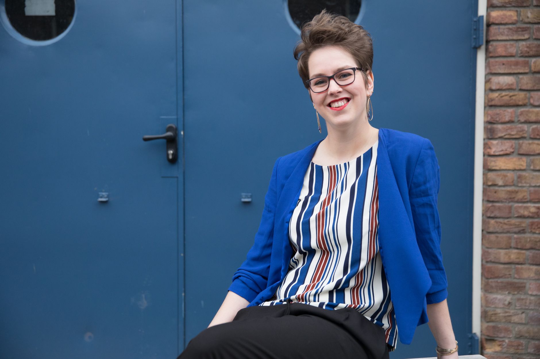 Smiling woman in a blue jacket sitting by a blue door, exuding confidence and style.
