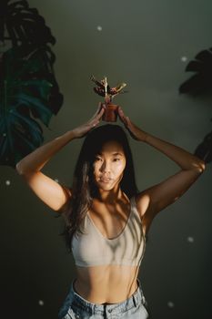 A woman in a tank top posing indoors with a plant on her head, illuminated by natural light.
