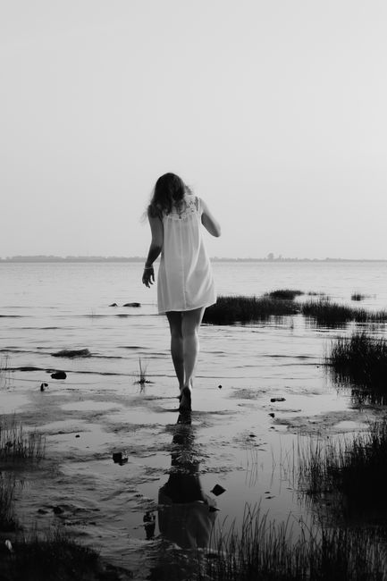 A woman in a white dress walks along a serene shoreline at dusk, captured in monochrome.