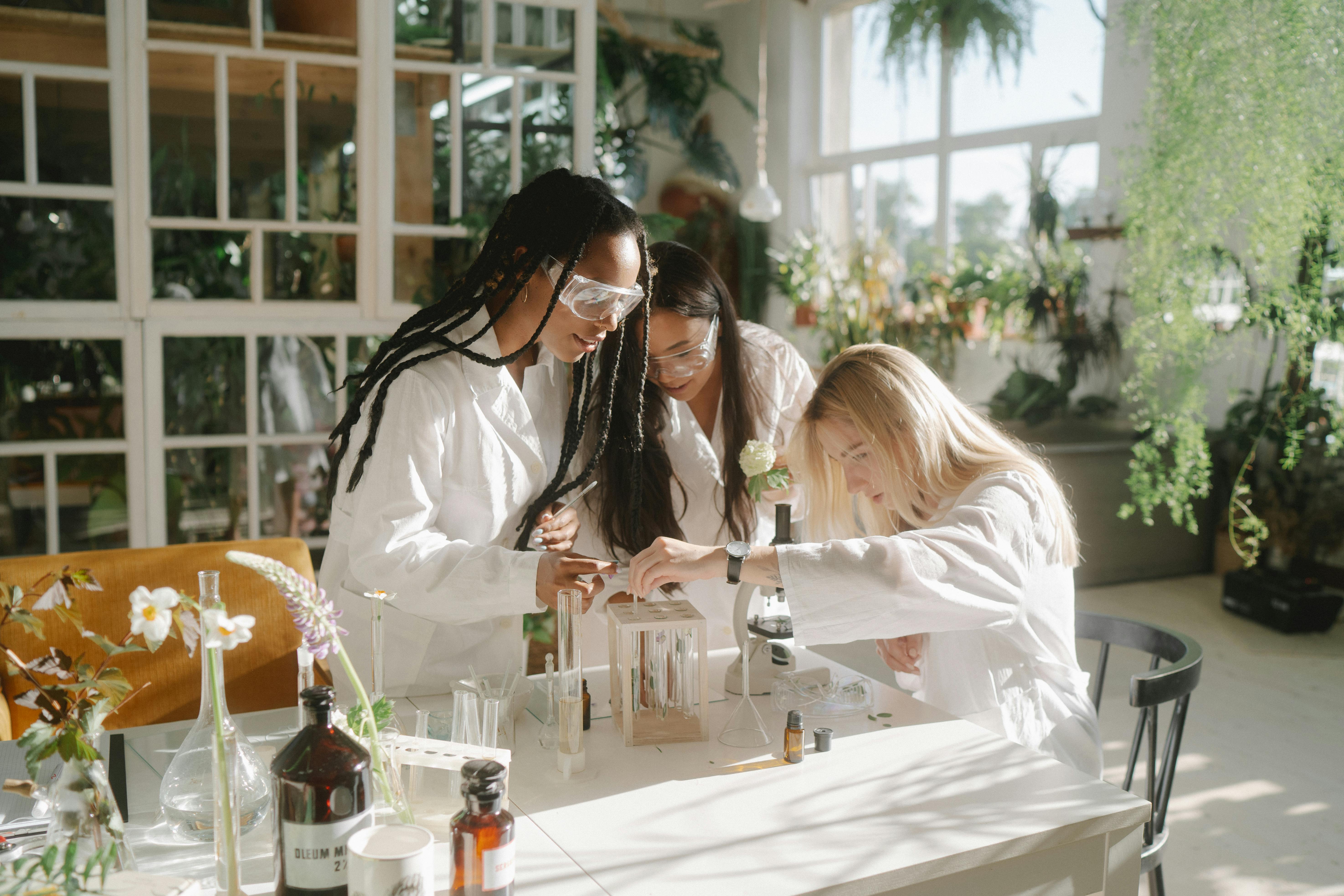 Three women in lab coats experimenting with plants and test tubes in a sunlit indoor lab.